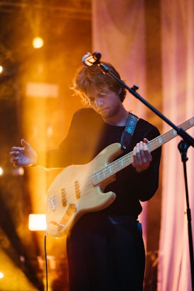 A musician is playing an electric bass guitar on stage, surrounded by warm, ambient lighting. The musician is focused, with one hand on the guitar neck and the other strumming the strings. The background features soft, blurred lighting effects and a microphone stand is positioned in front of the musician.