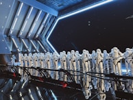 A large group of identically dressed armored figures standing in formation inside a futuristic, metallic room with angular architecture. The figures are wearing white armor with black details, resembling soldiers or guards. Blue lighting and reflections create a dramatic, sci-fi ambience.