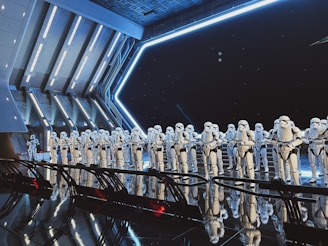 A large group of identically dressed armored figures standing in formation inside a futuristic, metallic room with angular architecture. The figures are wearing white armor with black details, resembling soldiers or guards. Blue lighting and reflections create a dramatic, sci-fi ambience.
