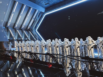 A large group of identically dressed armored figures standing in formation inside a futuristic, metallic room with angular architecture. The figures are wearing white armor with black details, resembling soldiers or guards. Blue lighting and reflections create a dramatic, sci-fi ambience.