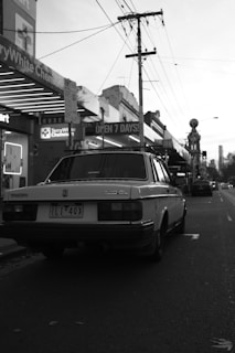 A classic car is parked on a dimly-lit street lined with shops. Power lines stretch above, and the ambiance is quiet with minimal activity. The storefront signs indicate a chemist and a supermarket open 7 days a week.