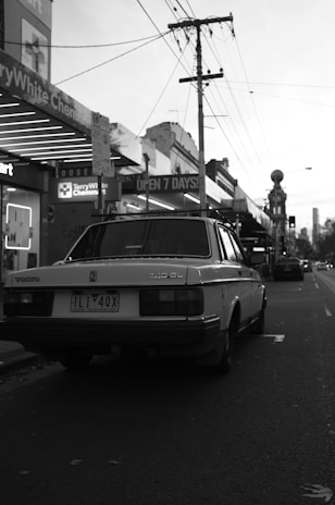 A classic car is parked on a dimly-lit street lined with shops. Power lines stretch above, and the ambiance is quiet with minimal activity. The storefront signs indicate a chemist and a supermarket open 7 days a week.