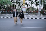 A man and a woman dressed in traditional attire walk hand in hand across a street. The background includes a monument with a gold and white column, green trees, and large red letters forming a word on the pavement. The couple seems happy and engaged in conversation.