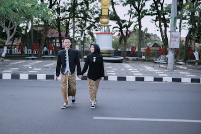 A man and a woman dressed in traditional attire walk hand in hand across a street. The background includes a monument with a gold and white column, green trees, and large red letters forming a word on the pavement. The couple seems happy and engaged in conversation.