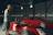 Two individuals stand near vintage racing cars inside a large, dimly lit garage. One person is wearing a yellow helmet and casual attire, examining the car closely. The other person is further back near another red car, adjusting their helmet and looking focused.