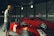 Two individuals stand near vintage racing cars inside a large, dimly lit garage. One person is wearing a yellow helmet and casual attire, examining the car closely. The other person is further back near another red car, adjusting their helmet and looking focused.