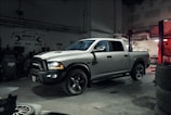 A silver pickup truck with black accents is parked in a dimly lit garage. The surroundings include various automotive equipment and tires. Overhead lighting casts shadows on the floor, contributing to a rugged atmosphere.