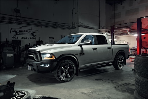 A silver pickup truck with black accents is parked in a dimly lit garage. The surroundings include various automotive equipment and tires. Overhead lighting casts shadows on the floor, contributing to a rugged atmosphere.
