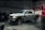 A silver pickup truck with black accents is parked in a dimly lit garage. The surroundings include various automotive equipment and tires. Overhead lighting casts shadows on the floor, contributing to a rugged atmosphere.