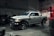 A silver pickup truck with black accents is parked in a dimly lit garage. The surroundings include various automotive equipment and tires. Overhead lighting casts shadows on the floor, contributing to a rugged atmosphere.