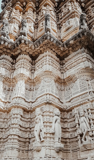 The image features an intricately carved wall of a temple or monument. The stone surface is adorned with detailed sculptures and ornate patterns, showcasing various figures and decorative elements. The architectural style appears to be traditional, with layered designs and significant attention to detail.
