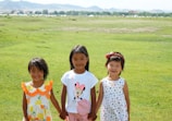 Three young children are standing on a vast green field. They are holding hands, smiling, and wearing colorful summer clothes. The background features open grassland extending towards a range of distant hills.