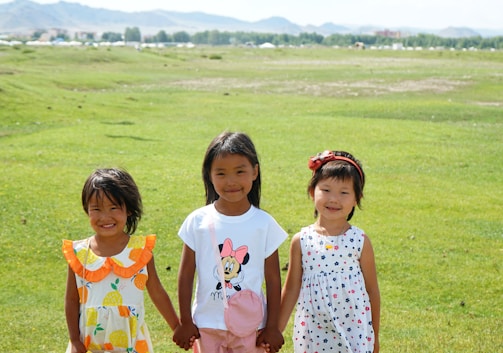 Three young children are standing on a vast green field. They are holding hands, smiling, and wearing colorful summer clothes. The background features open grassland extending towards a range of distant hills.