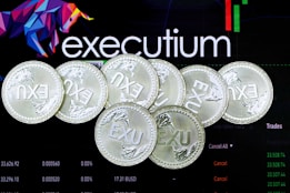 Several silver coins with the inscription 'EXU' are arranged in a pile. In the background, there is an electronic trading platform screen with the word 'executium', some graphs, and trading data.