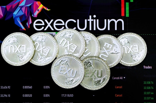 Several silver coins with the inscription 'EXU' are arranged in a pile. In the background, there is an electronic trading platform screen with the word 'executium', some graphs, and trading data.