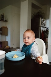 A young child seated in a high chair is holding a small piece of food. They are wearing a striped shirt and a blue bib, with a light blue bowl in front of them containing food remnants. The background includes a kitchen area with visible cabinets and a refrigerator.