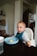 A young child seated in a high chair is holding a small piece of food. They are wearing a striped shirt and a blue bib, with a light blue bowl in front of them containing food remnants. The background includes a kitchen area with visible cabinets and a refrigerator.