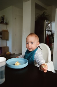 A young child seated in a high chair is holding a small piece of food. They are wearing a striped shirt and a blue bib, with a light blue bowl in front of them containing food remnants. The background includes a kitchen area with visible cabinets and a refrigerator.
