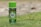 A can of Minyak Herba Sinergi is standing upright on grass. The can is predominantly green with decorative leaf patterns and has a logo featuring a bird. The text on the can reads 100 ml and indicates that it is a jamu product from HNI.