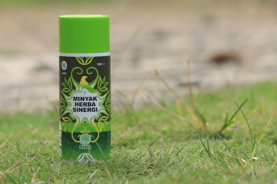 A can of Minyak Herba Sinergi is standing upright on grass. The can is predominantly green with decorative leaf patterns and has a logo featuring a bird. The text on the can reads 100 ml and indicates that it is a jamu product from HNI.