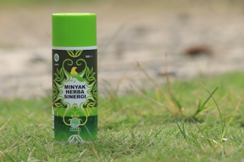 A can of Minyak Herba Sinergi is standing upright on grass. The can is predominantly green with decorative leaf patterns and has a logo featuring a bird. The text on the can reads 100 ml and indicates that it is a jamu product from HNI.