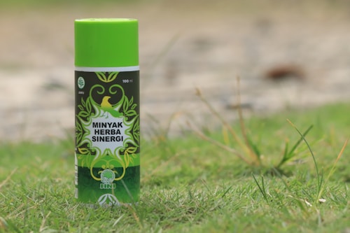 A can of Minyak Herba Sinergi is standing upright on grass. The can is predominantly green with decorative leaf patterns and has a logo featuring a bird. The text on the can reads 100 ml and indicates that it is a jamu product from HNI.