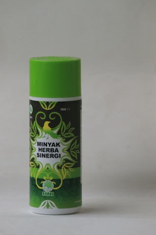 A cylindrical bottle with a bright green cap and label featuring decorative green leaves and a yellow bird. The label reads 'Minyak Herba Sinergi' in white text. The bottle appears to be 100 ml and has a logo with an abstract leaf design.