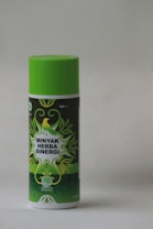 A cylindrical bottle with a bright green cap and label featuring decorative green leaves and a yellow bird. The label reads 'Minyak Herba Sinergi' in white text. The bottle appears to be 100 ml and has a logo with an abstract leaf design.