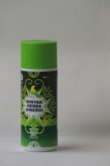 A cylindrical bottle with a bright green cap and label featuring decorative green leaves and a yellow bird. The label reads 'Minyak Herba Sinergi' in white text. The bottle appears to be 100 ml and has a logo with an abstract leaf design.