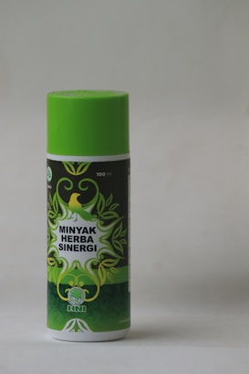 A cylindrical bottle with a bright green cap and label featuring decorative green leaves and a yellow bird. The label reads 'Minyak Herba Sinergi' in white text. The bottle appears to be 100 ml and has a logo with an abstract leaf design.