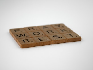 Wooden Scrabble tiles arranged to spell the words 'PRAY', 'WORK', 'REST' on a light grey background. The tiles have a natural wood finish with black letters and numbers indicating point values.