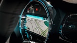 A car interior focusing on the dashboard with a built-in GPS navigation system. The screen displays a map with streets highlighted in green and gray. The dashboard has a modern design with illuminated blue lights outlining the circular screen.