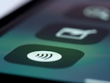 A close-up of a mobile device screen displaying a contactless payment icon in the form of a series of curved lines. The background includes blurred app icons with dark and muted tones.