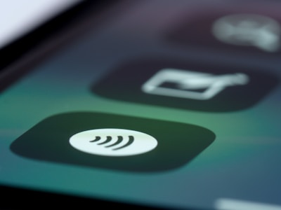 A close-up of a mobile device screen displaying a contactless payment icon in the form of a series of curved lines. The background includes blurred app icons with dark and muted tones.