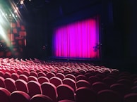 A theater setting with rows of red cushioned seats facing a stage. The stage is framed by a bright pink curtain, and overhead lights illuminate parts of the space, adding a dramatic effect.
