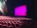 A theater setting with rows of red cushioned seats facing a stage. The stage is framed by a bright pink curtain, and overhead lights illuminate parts of the space, adding a dramatic effect.