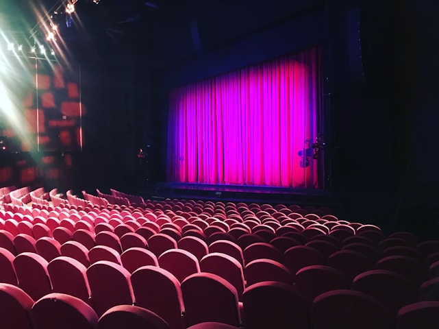 A theater setting with rows of red cushioned seats facing a stage. The stage is framed by a bright pink curtain, and overhead lights illuminate parts of the space, adding a dramatic effect.