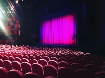 A theater setting with rows of red cushioned seats facing a stage. The stage is framed by a bright pink curtain, and overhead lights illuminate parts of the space, adding a dramatic effect.