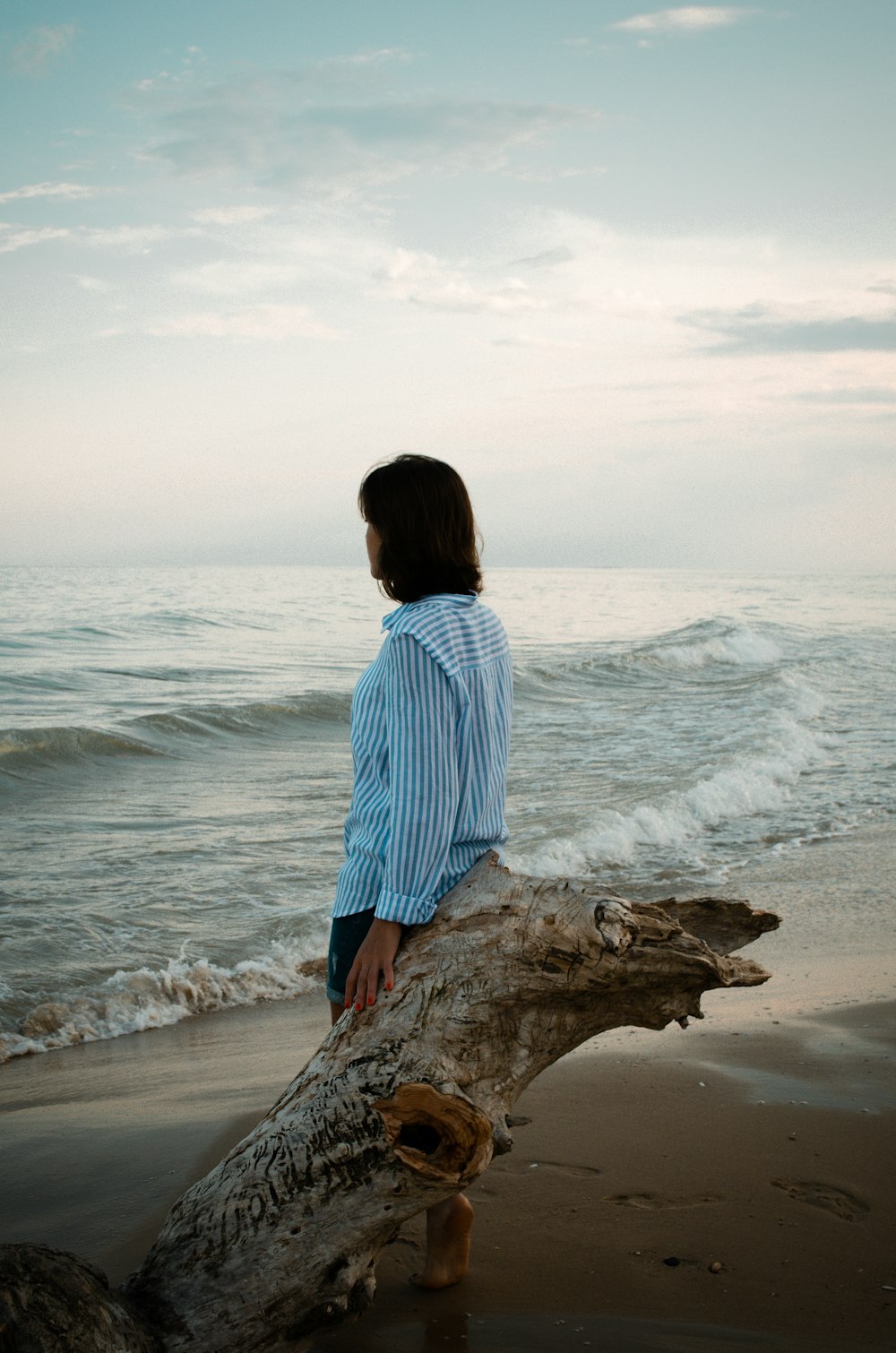 woman in blue and white stripe shirt sitting on brown tree trunk on beach during daytime