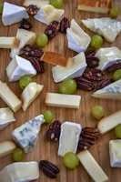 An assortment of various types of cheeses, including brie and blue cheese, arranged on a wooden surface. Scattered among the cheeses are whole pecan nuts and green grapes.