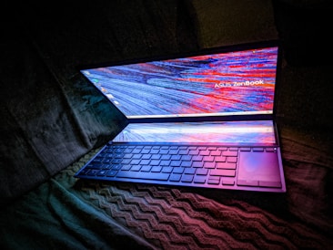 A laptop with a glowing dual-screen display, featuring vibrant and colorful abstract patterns. The keyboard is illuminated with a soft blue and purple hue, contrasting against a dark background.