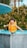 A child wearing a bright orange flotation vest stands at the edge of a pool, appearing surprised or excited. The water is a vivid blue, and there are green bushes in the background.