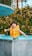 A child wearing a bright orange flotation vest stands at the edge of a pool, appearing surprised or excited. The water is a vivid blue, and there are green bushes in the background.