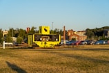 A small outdoor cafe with a vibrant yellow and black exterior sits in an open grassy area. It features a serving window with beverages and snacks displayed. Several picnic tables are arranged around the cafe, providing seating for visitors. In the background, a parking lot is filled with parked cars, and beyond that, a few industrial-style brick buildings and lines of trees are visible.