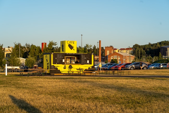 A small outdoor cafe with a vibrant yellow and black exterior sits in an open grassy area. It features a serving window with beverages and snacks displayed. Several picnic tables are arranged around the cafe, providing seating for visitors. In the background, a parking lot is filled with parked cars, and beyond that, a few industrial-style brick buildings and lines of trees are visible.