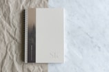 A spiral-bound notebook rests on a surface that transitions from crumpled brown paper to smooth white marble. The notebook's cover is a muted beige with a subtle design featuring a tree, and the initials 'SK' are embossed at the bottom corner.