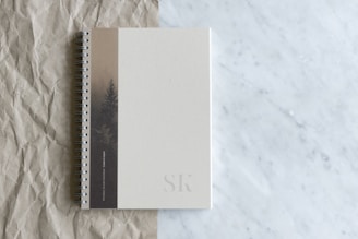 A spiral-bound notebook rests on a surface that transitions from crumpled brown paper to smooth white marble. The notebook's cover is a muted beige with a subtle design featuring a tree, and the initials 'SK' are embossed at the bottom corner.