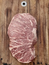 A large piece of marbled raw meat placed on a wooden cutting board. The fat creates intricate patterns across the meat, and there's a metal hole at the top of the board.