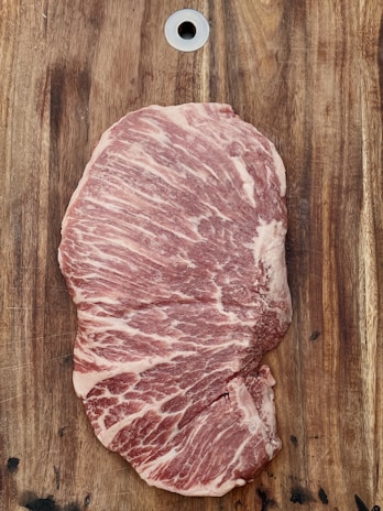 A large piece of marbled raw meat placed on a wooden cutting board. The fat creates intricate patterns across the meat, and there's a metal hole at the top of the board.