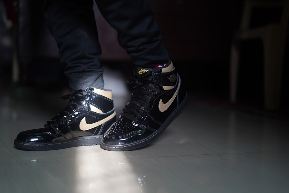 A pair of black and gold high-top sneakers with a shiny finish is being worn indoors. The lighting highlights the glossy texture of the shoes. The person is wearing dark pants, and the background includes a dimly lit interior with part of a chair visible.